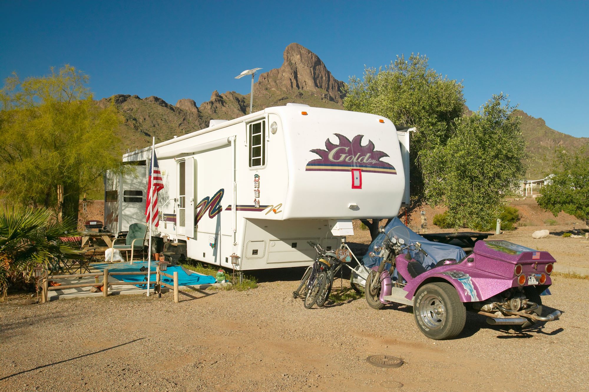 An off-road RV parked in a desert mountain area with an American flag, set of bikes, and an ATV