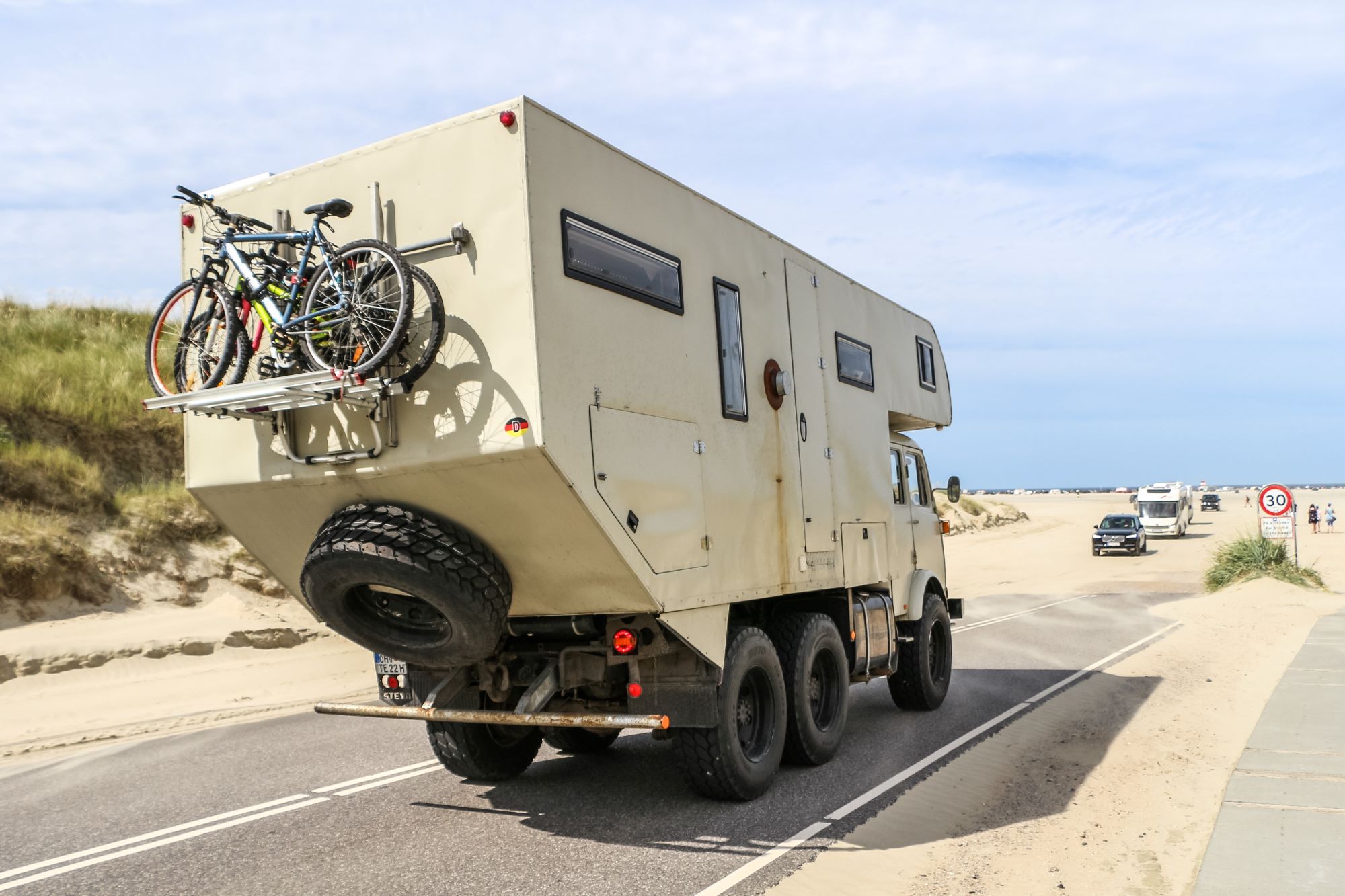 An RV Camper driving down a desert highway with bikes and a spare tire on its back