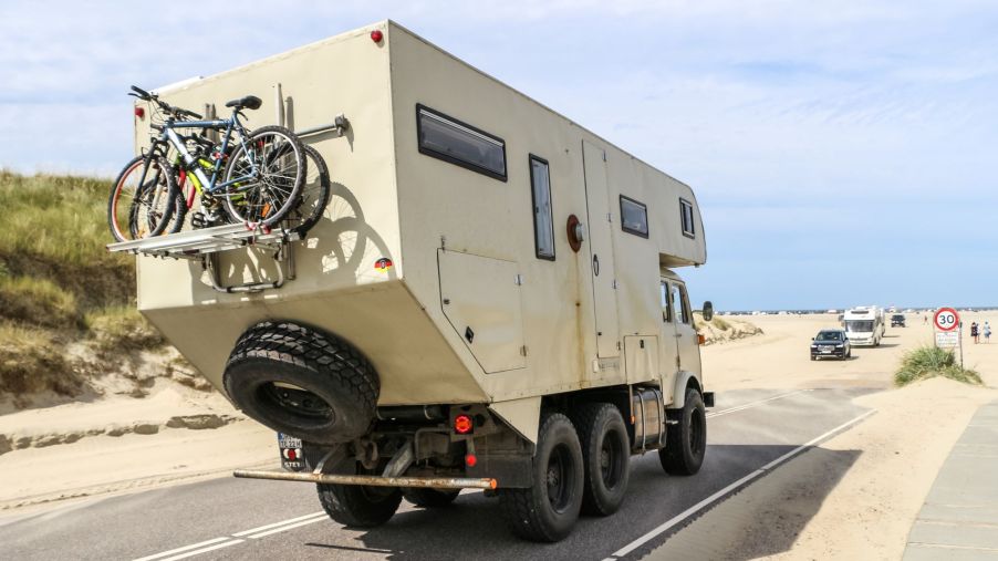 An RV Camper driving down a desert highway with bikes and a spare tire on its back