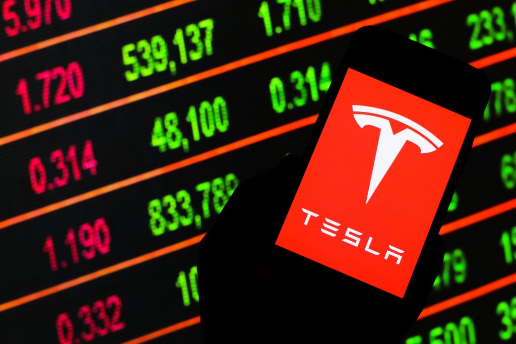 A Tesla logo displayed on a smartphone in front of lighted stock ticker board