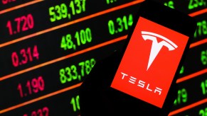 A Tesla logo displayed on a smartphone in front of lighted stock ticker board