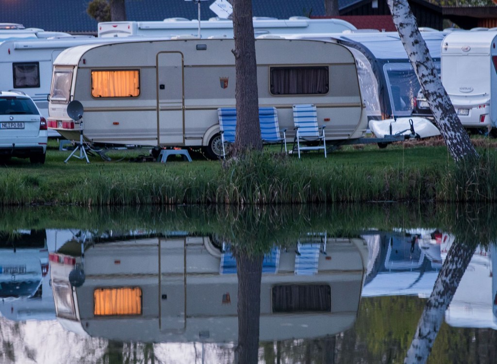 A cream and gray travel trailer with a single tree in the middle front, parked beyond a pond where the reflection of the RV can be seen. There are two darker blue and one lighter blue lawn chair in front of the travel trailer. There are other cars and travel trailers in the background.