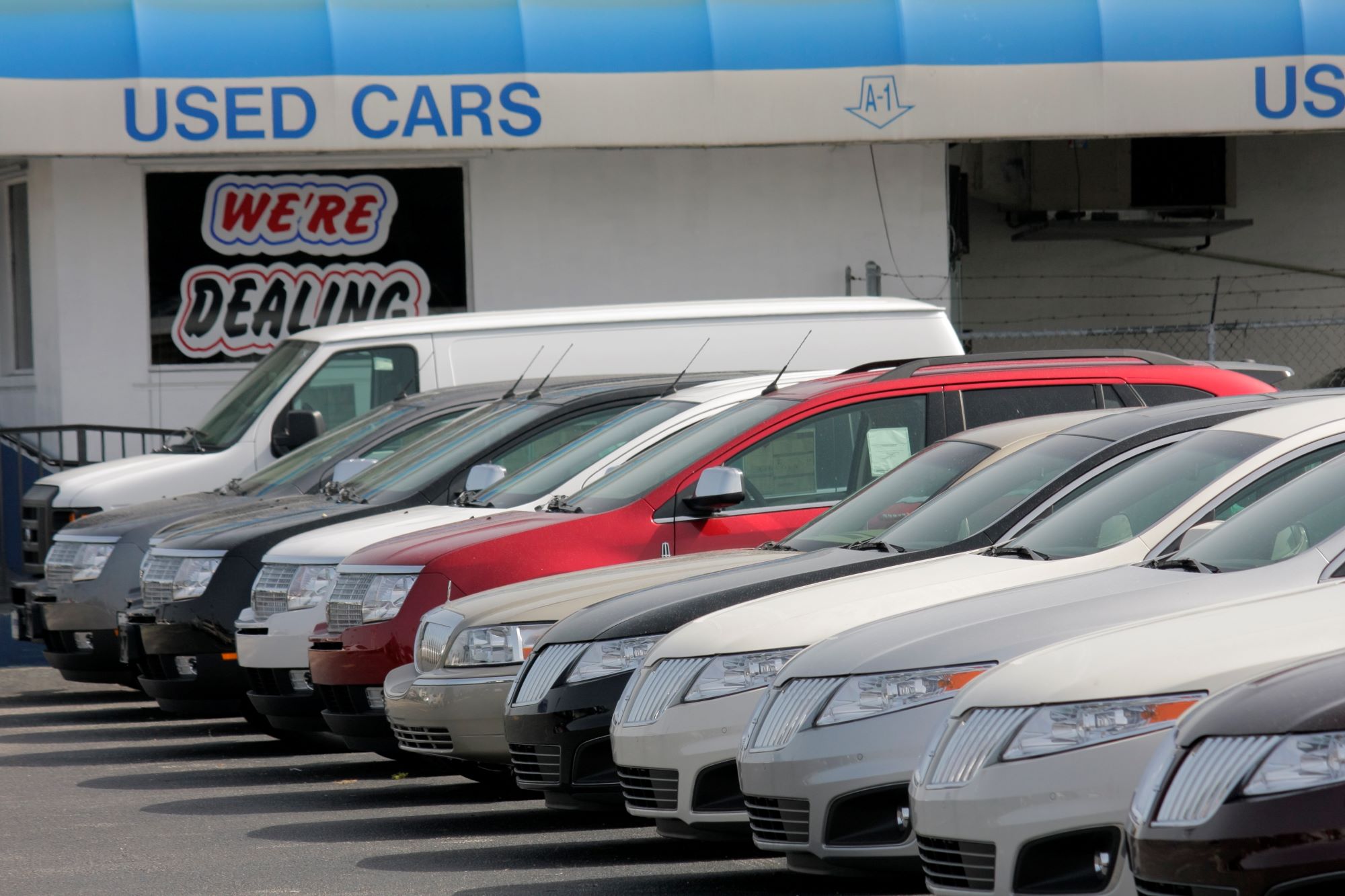 A picture of a used car lot similar to a buy here pay here with numerous cars of various colors in front of a small white building with a blue overhang that says "used cars" with a sticker in a blacked out window that says "We're Dealing."
