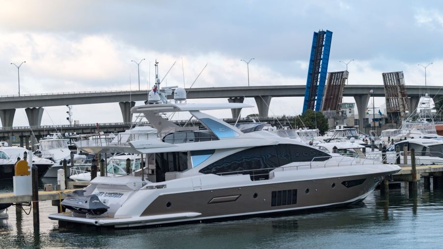 A smaller white and grey yacht parked in a marina with numerous yachts and a bridge in the background