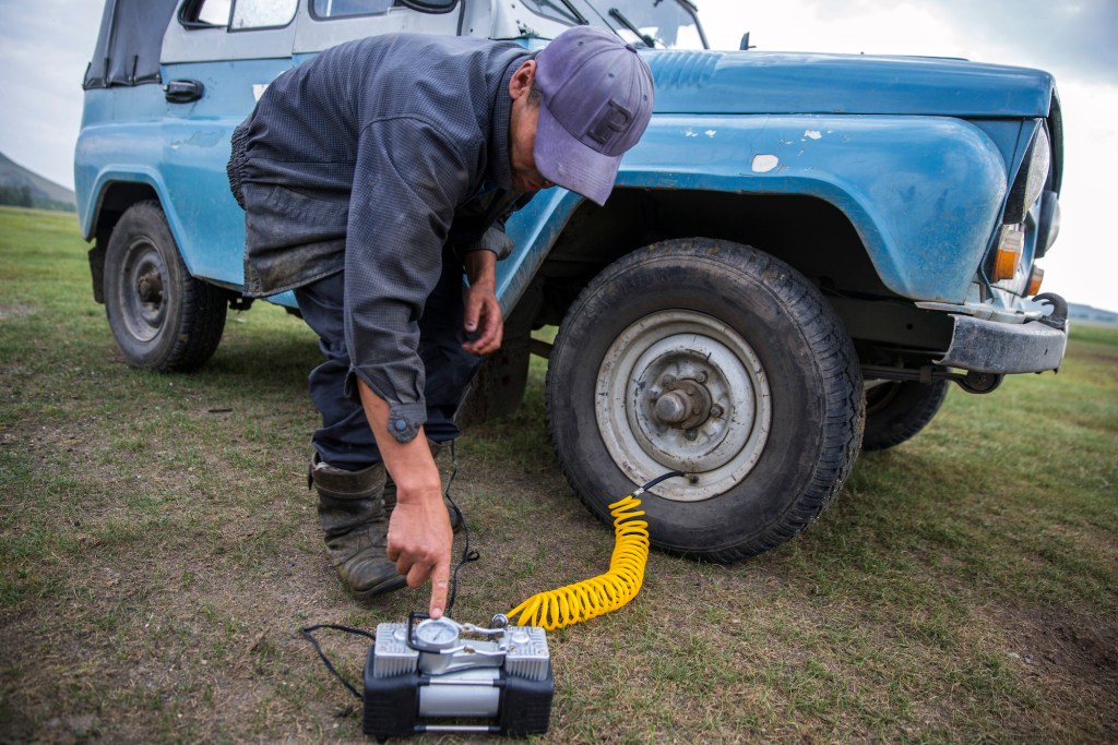 A man turns on a battery-powered air compressor whose hose is attached to his SUV's right front tire