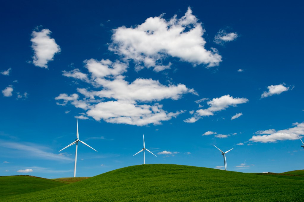 A clear blue sky with white clouds above wind turbines on green grass-covered hills