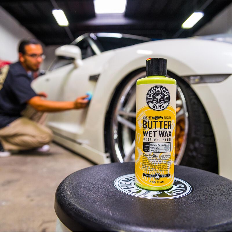Chemical Guys Butter Wet car wax sits on a stool while a man polishes a white Nissan GTR in the background