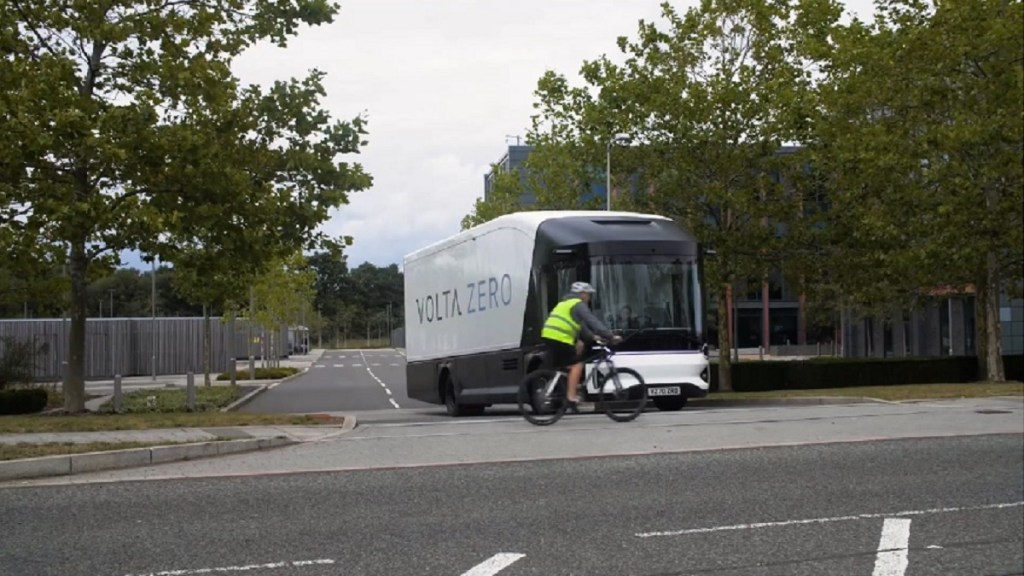 A Volta Zero truck pulls out of a side street as a cyclist pedals past it.