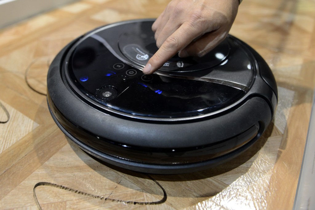 A person's finger presses a button on a robot vaccum cleaner sitting on linoleum