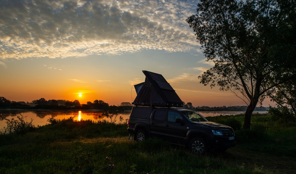 An off-road vehicle with an open roof tent stands on the banks of the Elbe river at sunrise.