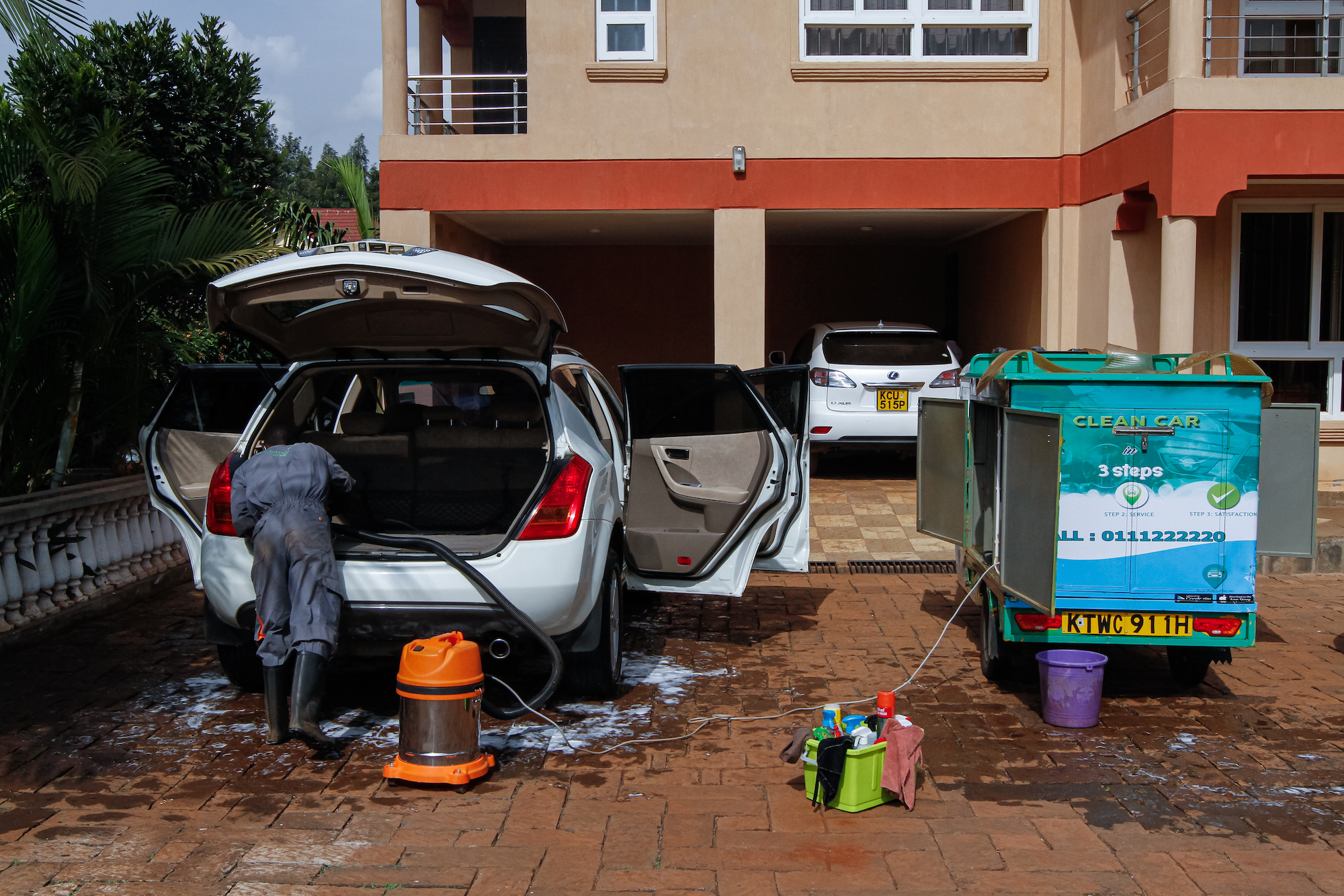 A mobile car wash worker uses a shop vac to vacuum the back of an SUV in a customer's front yard