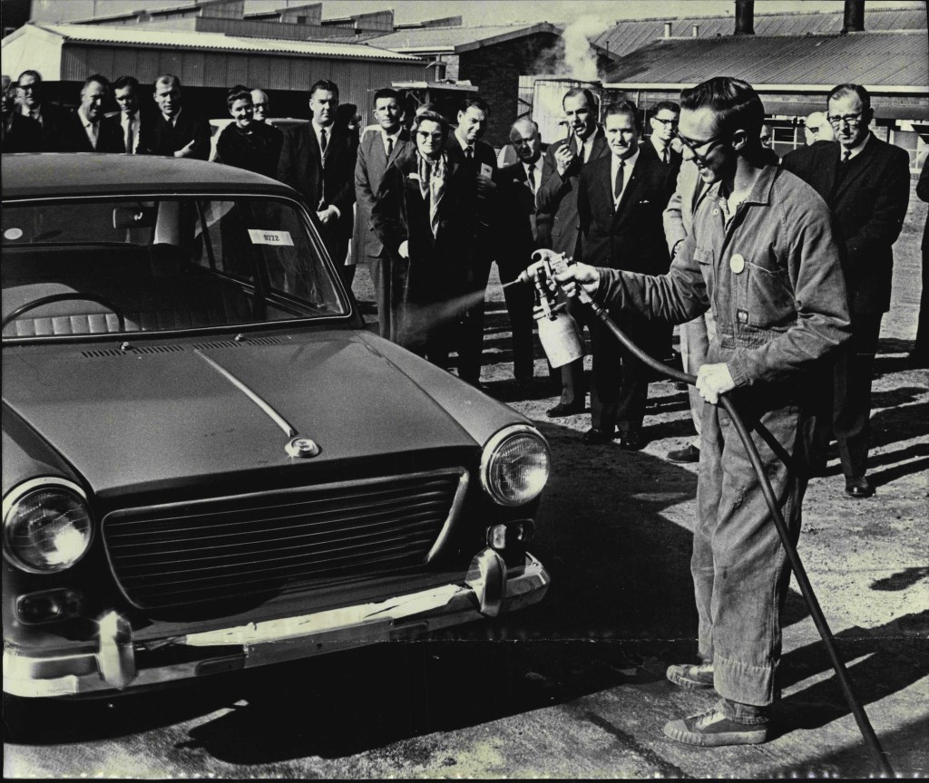 Businesspeople watch a worker use a spray car wax on a vehicle in 1964