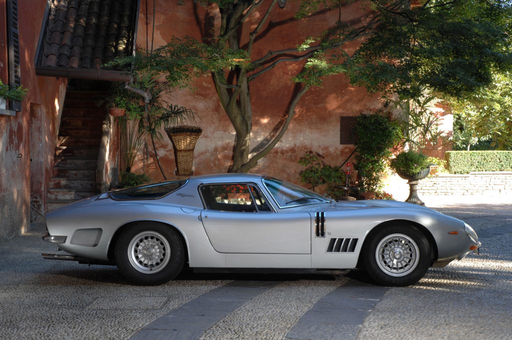 The side view of a silver 1966 Bizzarrini 5300 GT Strada parked by a villa