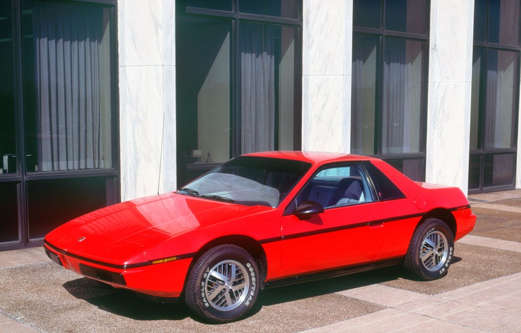 A red 1983 Pontiac Fiero in front of white pillars and a brown building lined with windows.
