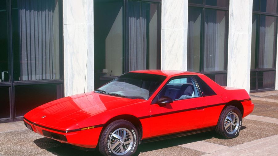 A red 1983 Pontiac Fiero in front of white pillars and a brown building lined with windows.
