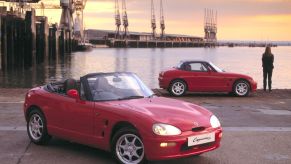Two red 1991 Suzuki Cappucinos--one with its roof up, one with its roof down--next to a harbor