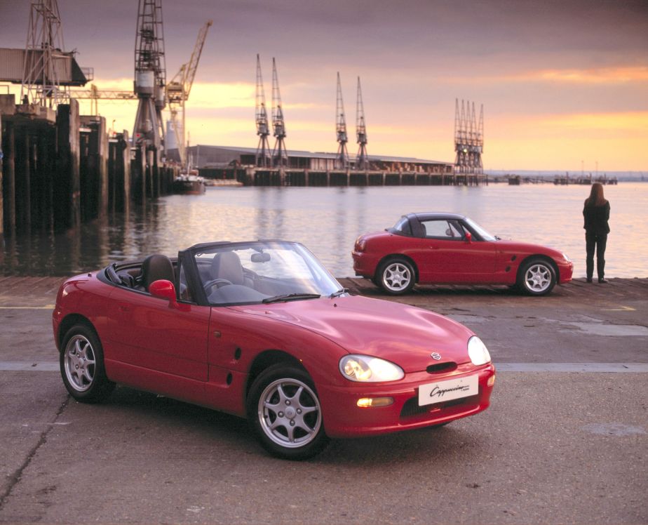 Two red 1991 Suzuki Cappucinos--one with its roof up, one with its roof down--next to a harbor