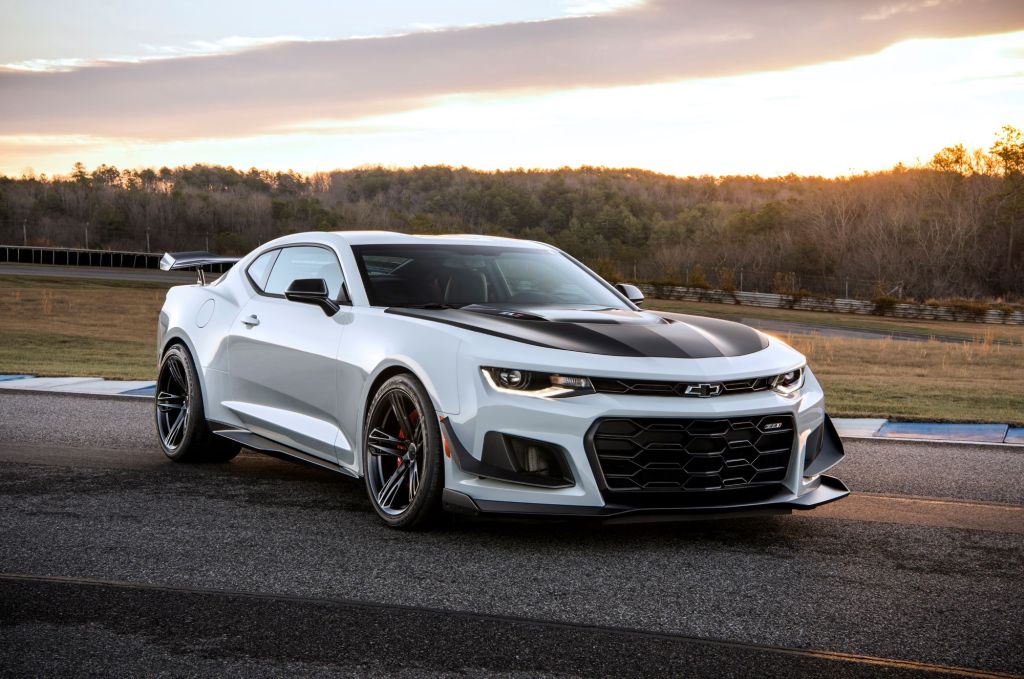 A silver Camaro with black racing stripes on the hood sitting on blacktop in a wooded area as the background.