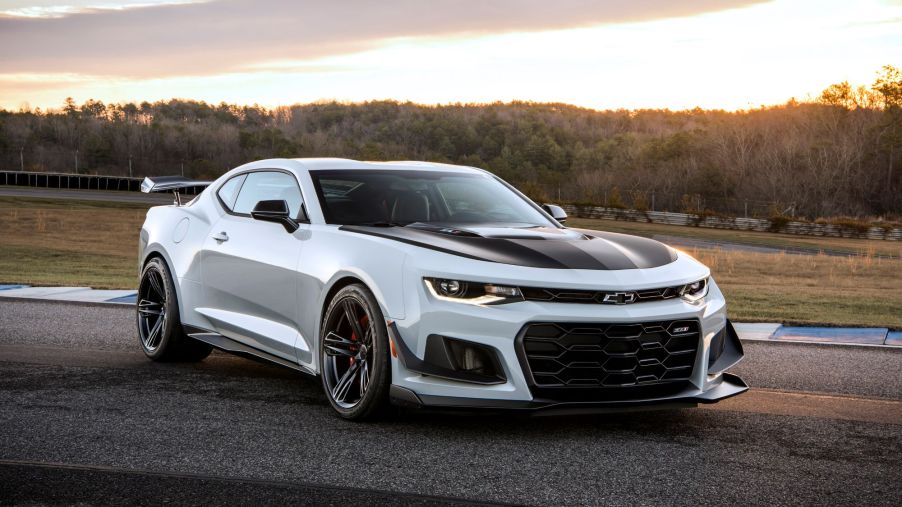 A silver Camaro with black racing stripes on the hood sitting on blacktop in a wooded area as the background.