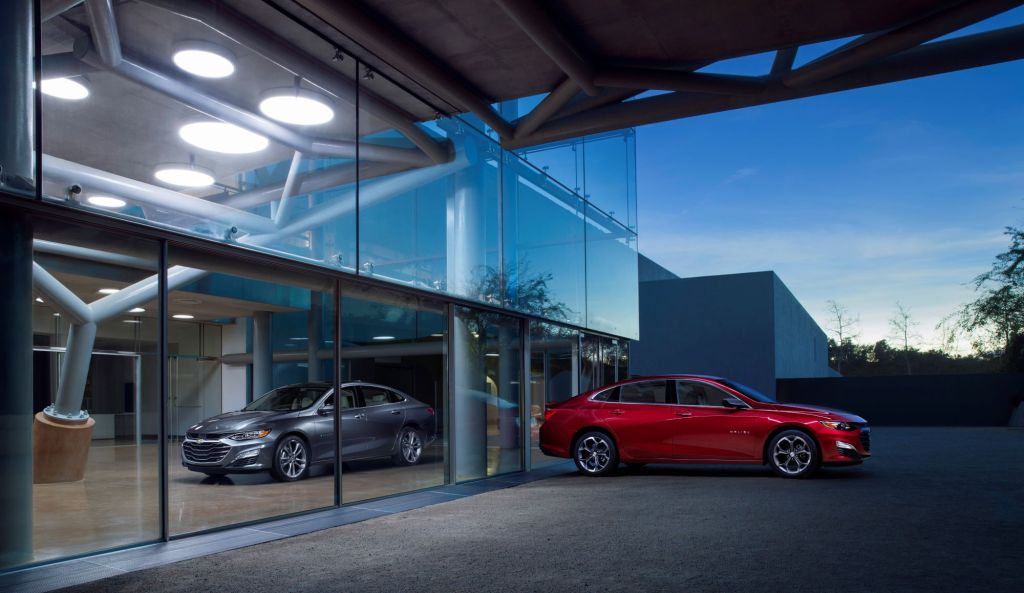 A red 2020 Chevrolet Malibu sits outside at twilight in front of a building with glass windows with a grey 2020 Chevrolet Malibu sitting behind the red one inside.