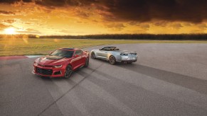 A red 2021 Chevy Camaro ZL1 coupe and silver ZL1 convertible parked on asphalt overlooking a large expanse of grass as the sun hangs low on the horizon