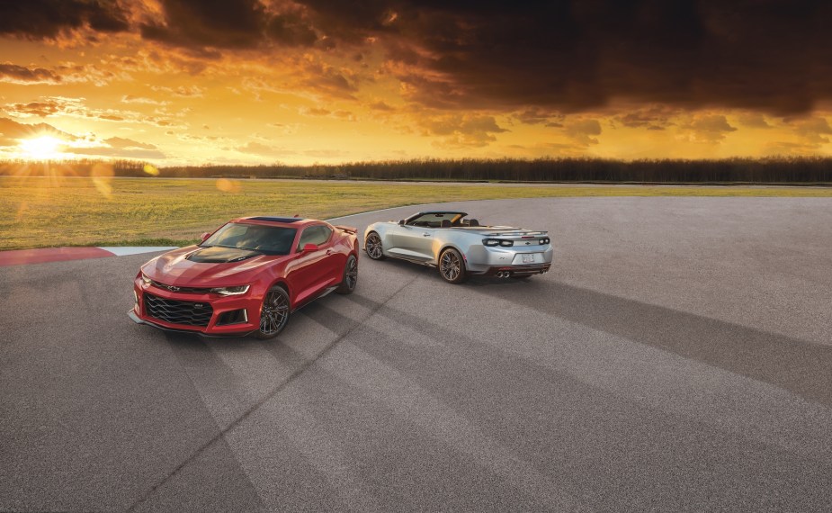 A red 2021 Chevy Camaro ZL1 coupe and silver ZL1 convertible parked on asphalt overlooking a large expanse of grass as the sun hangs low on the horizon