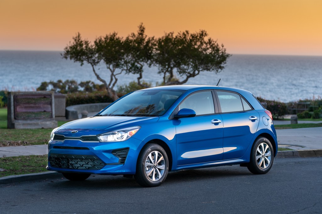 A blue metallic 2021 Kia Rio 5-Door Hatchback parked on a street overlooking a body of water at sunset
