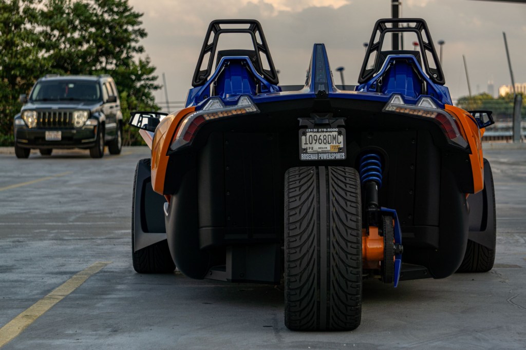 The rear view of an orange-and-blue 2021 Polaris Slingshot R in a parking lot with a Jeep SUV in the background