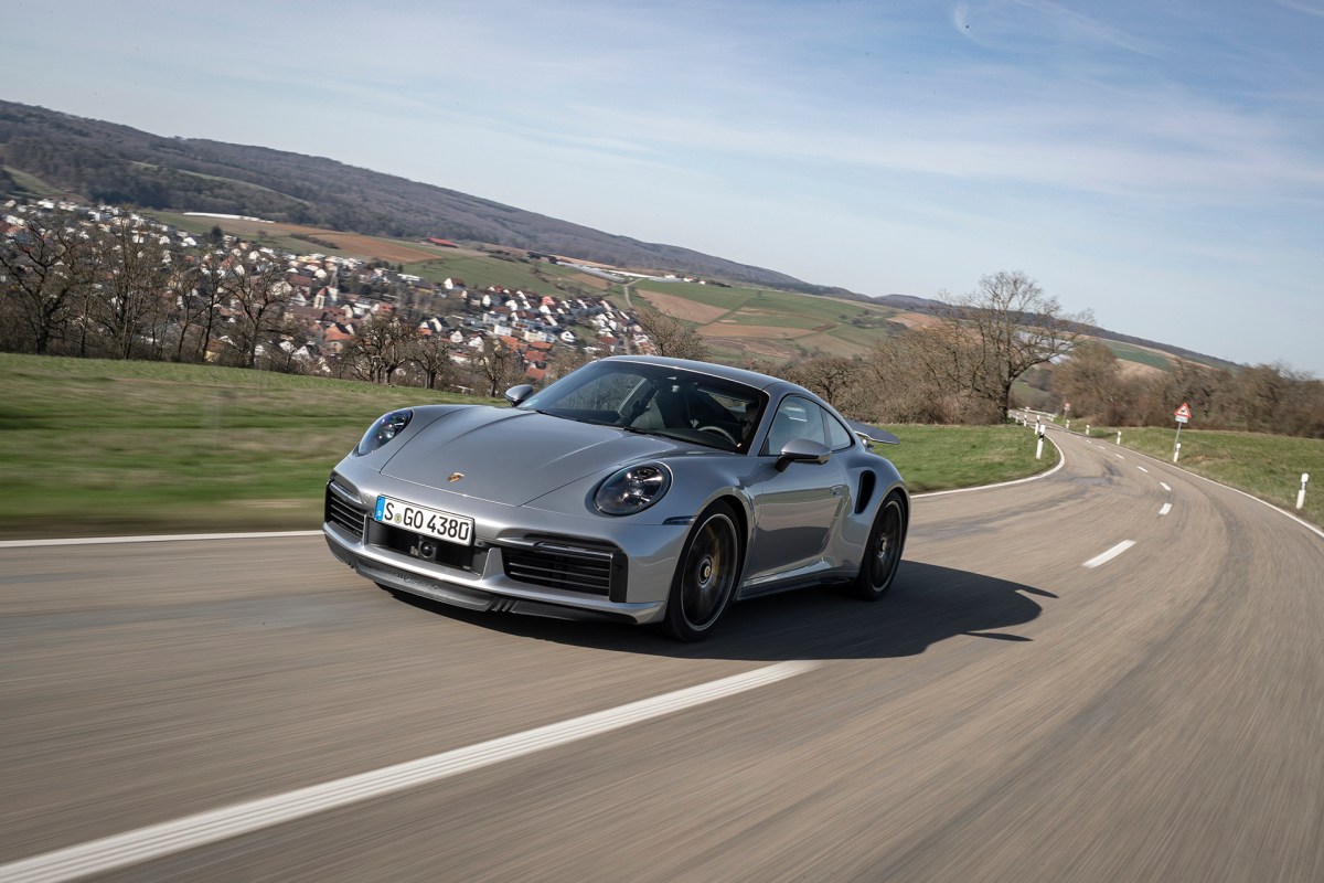 A sliver 2021 Porsche 911 Turbo S racing down an empty backroad on a country side.