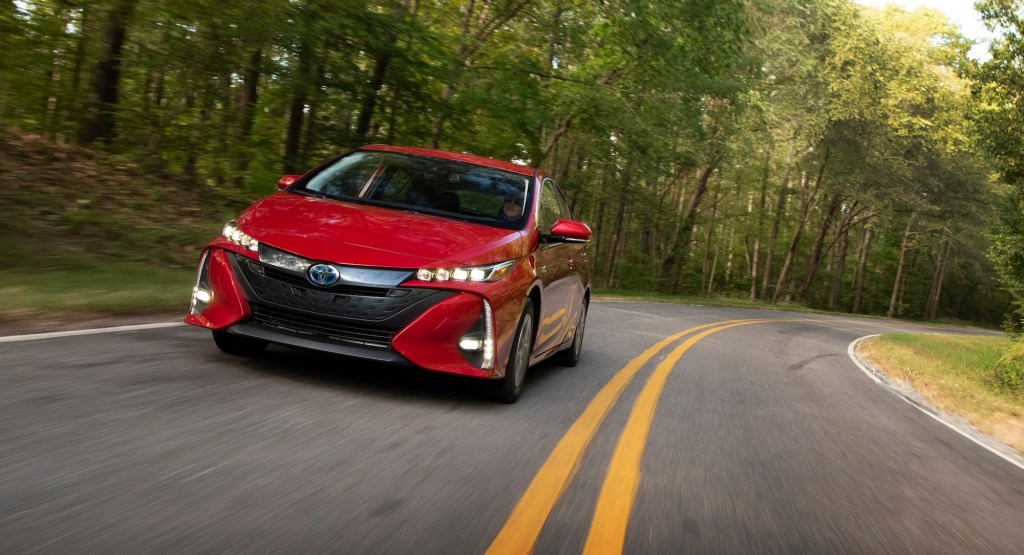 A red 2021 Toyota Prius Prime travels on a two-lane highway through sun-dappled trees