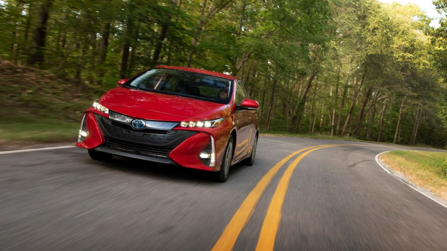 A red 2021 Toyota Prius Prime travels on a two-lane highway through sun-dappled trees