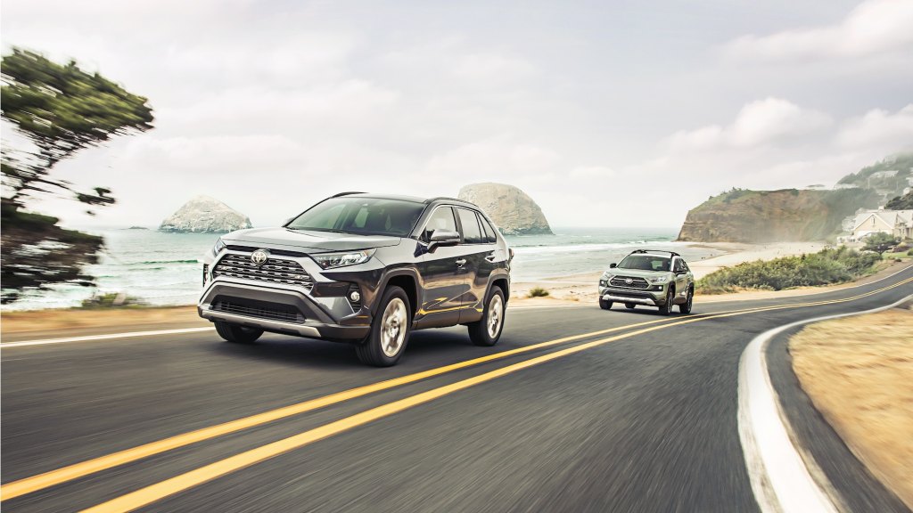 A dark-gray metallic 2021 Toyota RAV4 Limited AWD compact SUV traveling in front of a gray 2021 Toyota RAV4 Adventure on a two-lane highway overlooking cliffs and an ocean