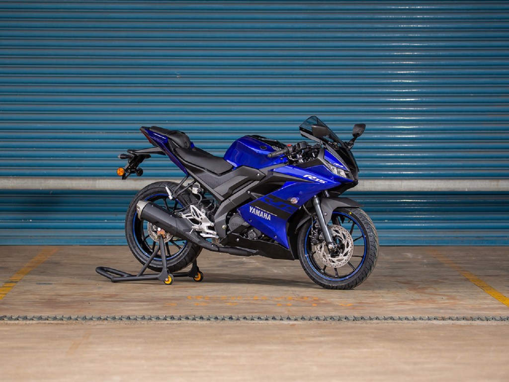 A blue-and-black 2021 Yamaha YZF-R15 on a rear-wheel stand by a turquoise garage