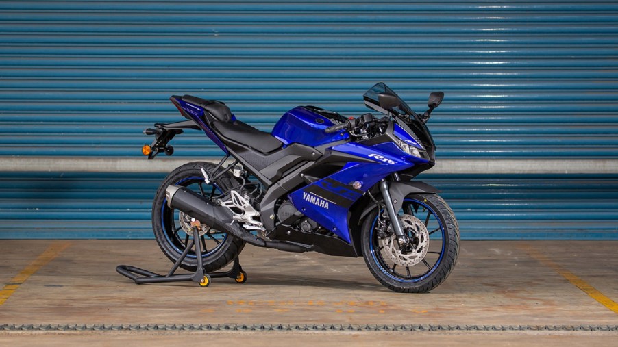 A blue-and-black 2021 Yamaha YZF-R15 on a rear-wheel stand by a turquoise garage