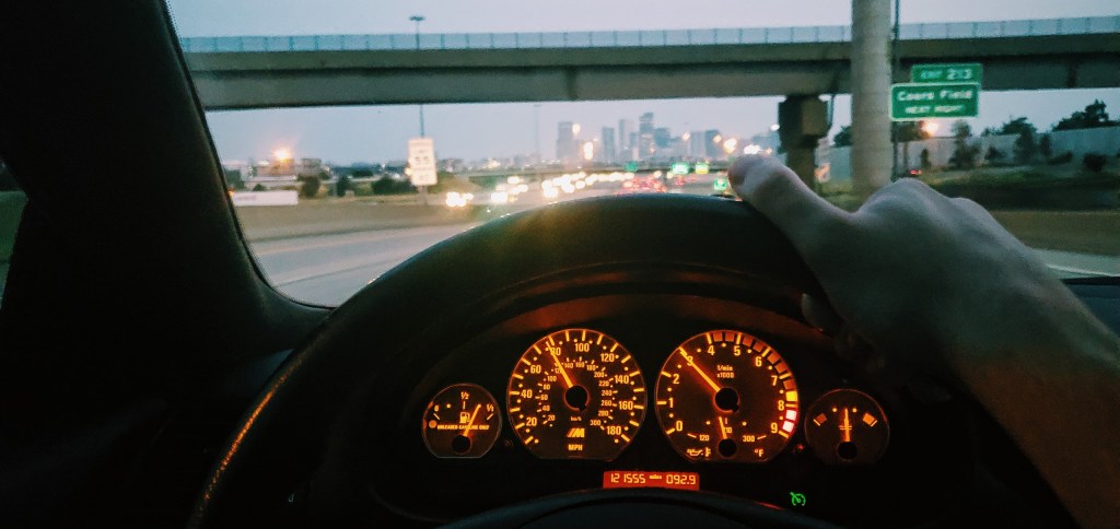The amber gauge cluster of a 2004 BMW M3 at dusk with the city in the background