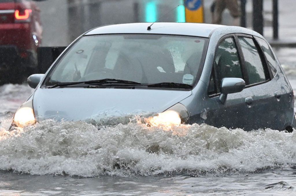 A blue hatchback driving through flood waters that come almost over the headlights. 