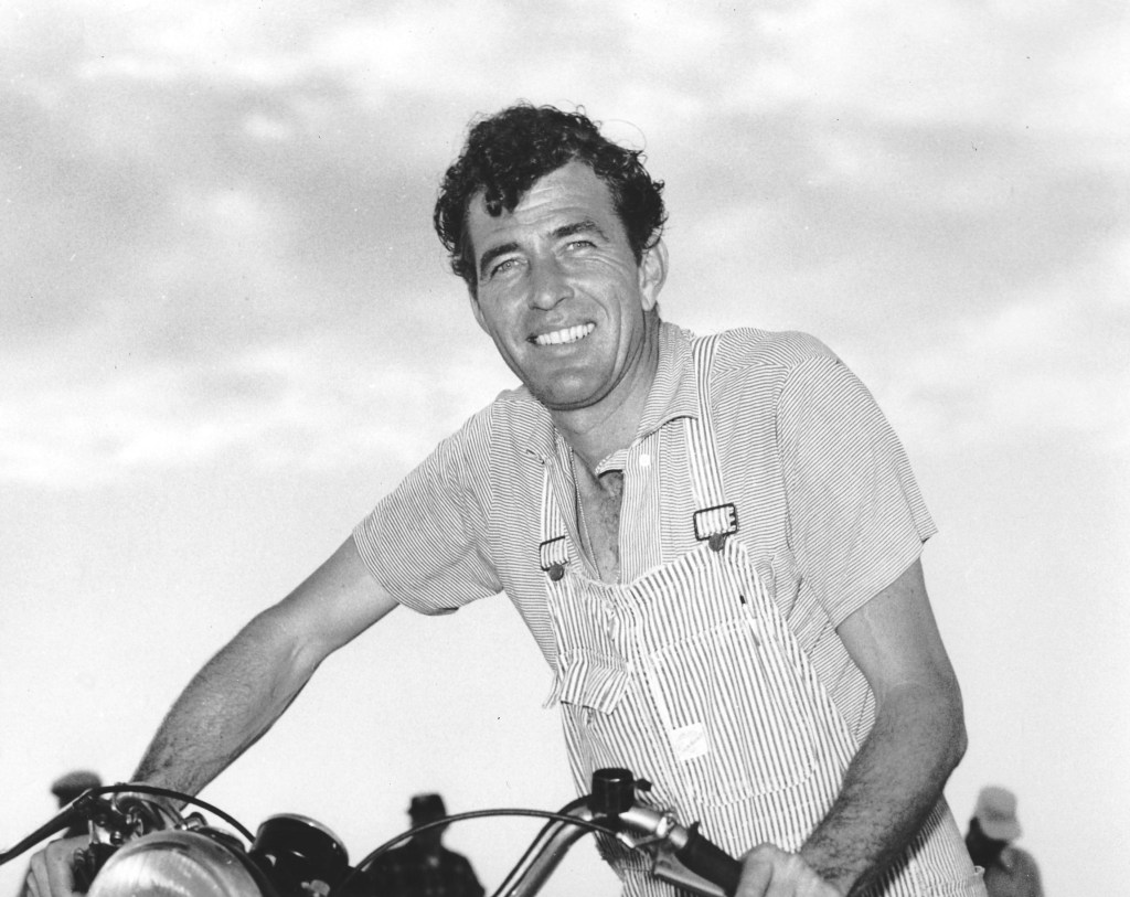 Carroll Shelby, the father of the Shelby Cobra, wearing his overalls while pushing a motorcycle. 