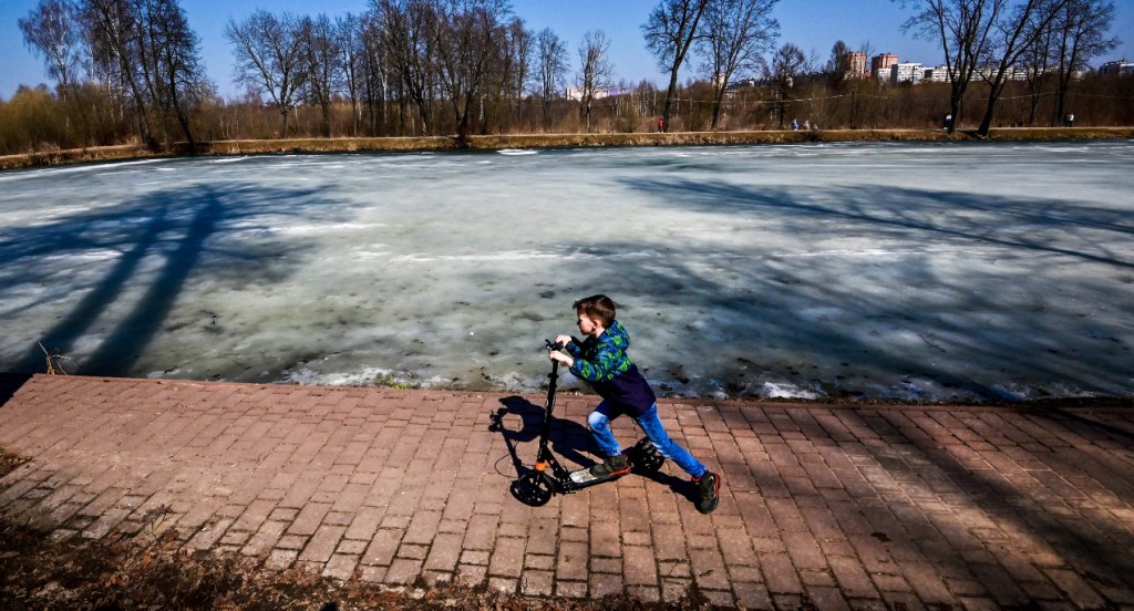 A boy rides a scooter as a frozen pond is seen in the background.