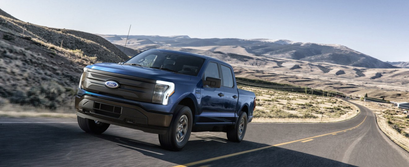 A blue 2022 Ford F-150 Lightning Pro driving down an empty street in the wilderness