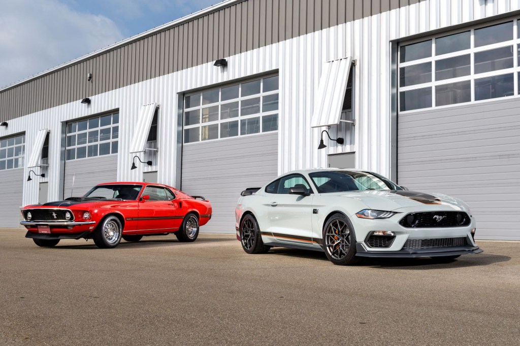 A white 2021 Mustang Mach 1 sitting in front of an older orange model in front of a metal garage style building. 