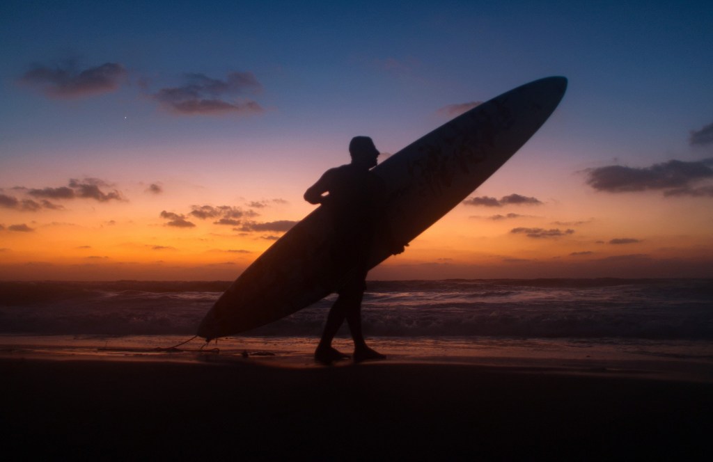 A man carries a surfboard on the beach, silhouetted by the sunset