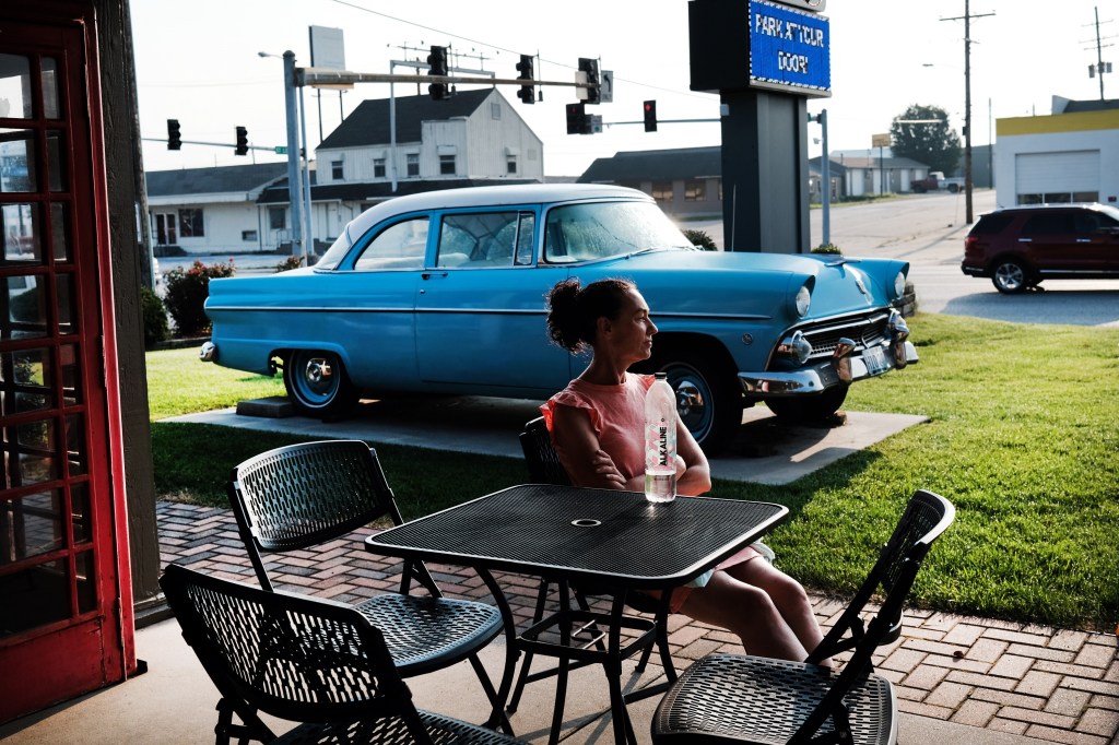 A woman sits on a patio with a 50's-era car in the background