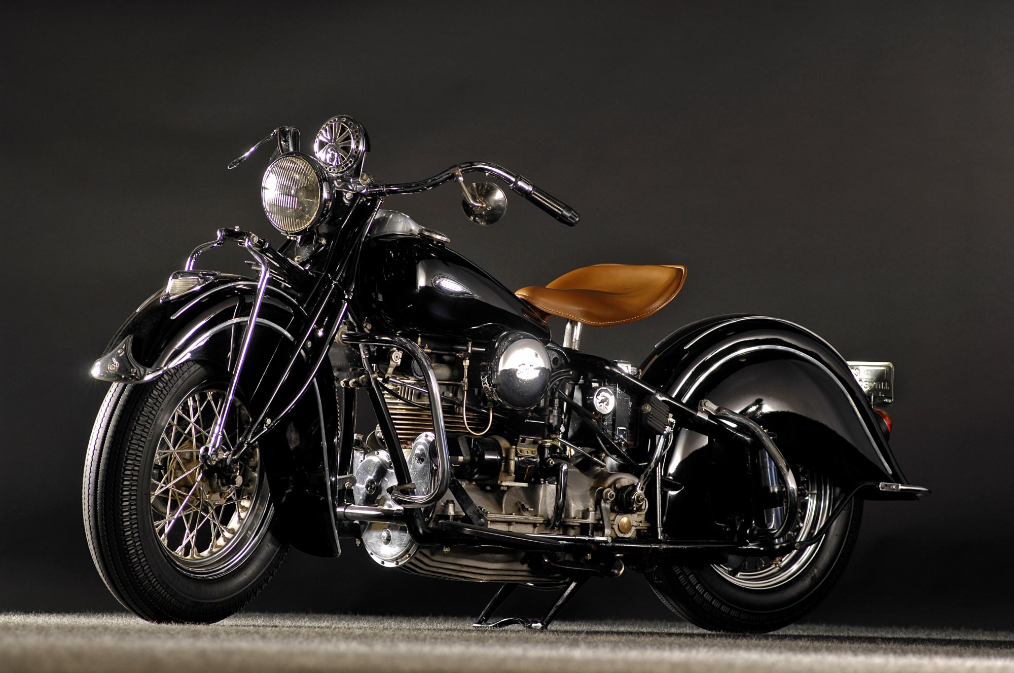 An old black Indian Motorcycle with a tan seat against a dark grey background.