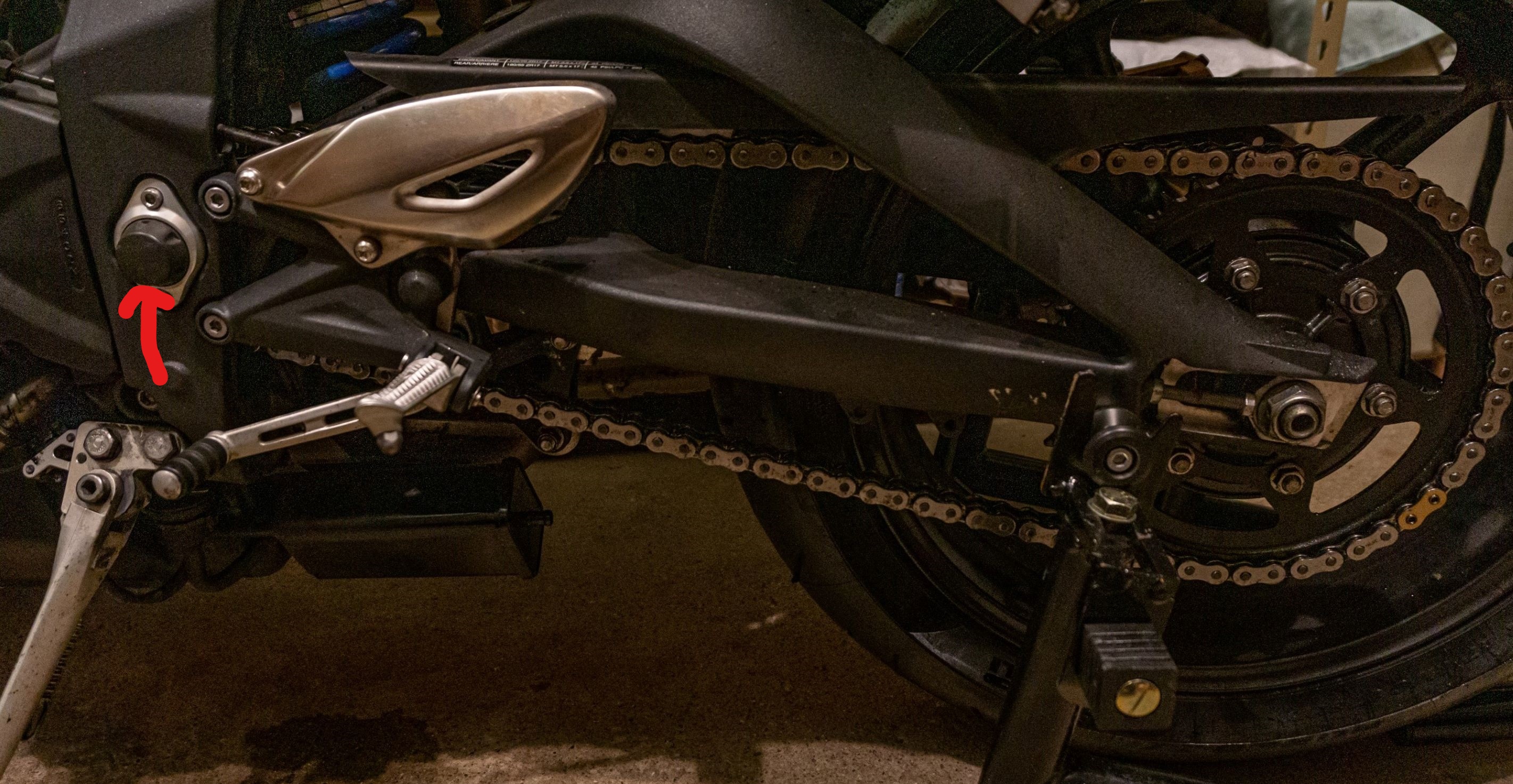 A 2012 Triumph Street Triple R motorcycle's chain and rear sprocket with its swingarm pivot indicated by a red arrow