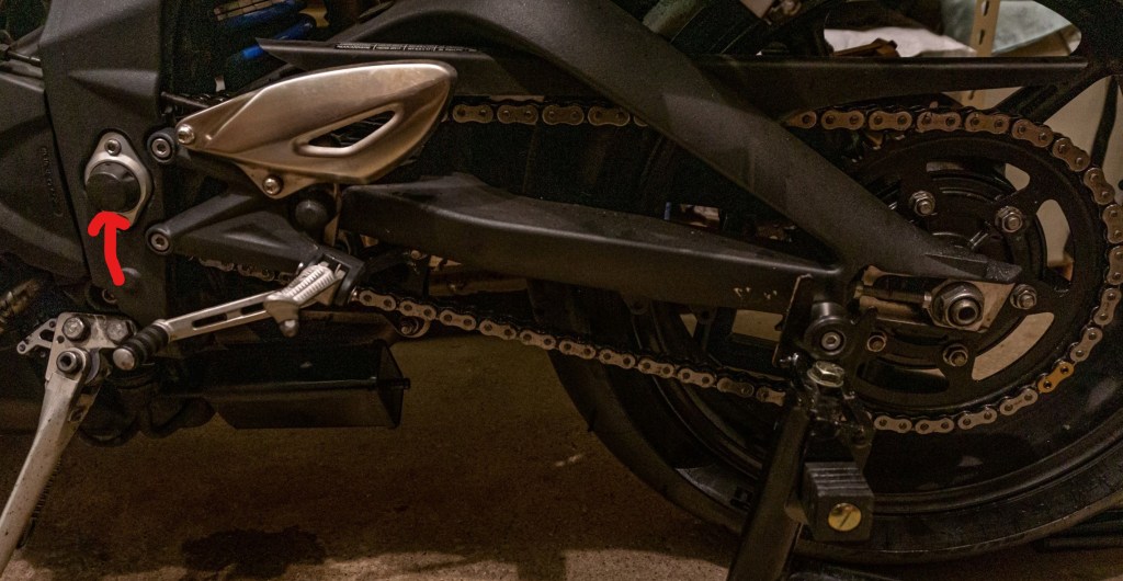 A 2012 Triumph Street Triple R motorcycle's chain and rear sprocket with its swingarm pivot indicated by a red arrow