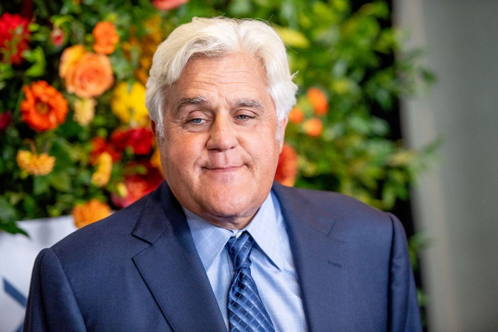 Jay Leno wearing a blue suit with a light blue shirt and a dark stripped blue tie in front of a flowered background. 