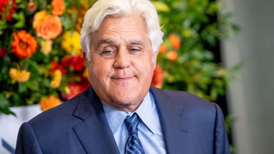 Jay Leno wearing a blue suit with a light blue shirt and a dark stripped blue tie in front of a flowered background.