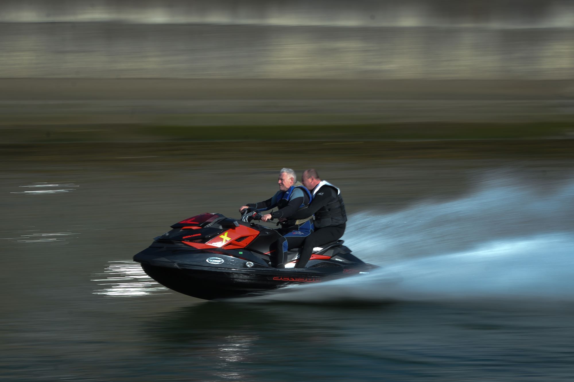 2 men riding on a black and red jet ski in a large body of water wearing black jackets and pants.