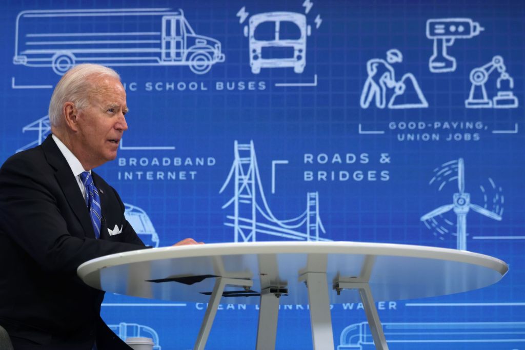 Joe Biden sitting at a white table in a chair dressed in a suit and tie in front of a blue background with engineering sketches of various things, such as road and bridges, wind turbines, electric vehicles.