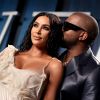 Kim Kardashian West is dressed in an ivory dress with Kanye West in a black shiny suit jacket in front of a blue, black and white background.